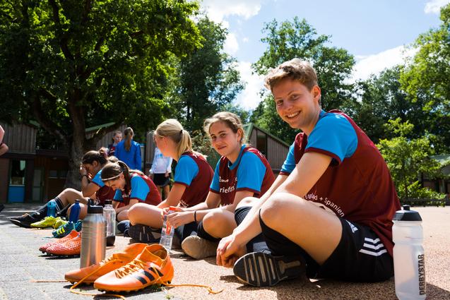 Eine Gruppe junger Fußballspielerinnen sitzt entspannt auf dem Boden, während sie ihre Schuhe ausziehen und sich unterhalten.