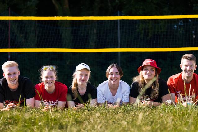 Sechs Personen liegen im Gras vor einem Beachvolleyballfeld mit gelben Netzen und Bäumen im Hintergrund.