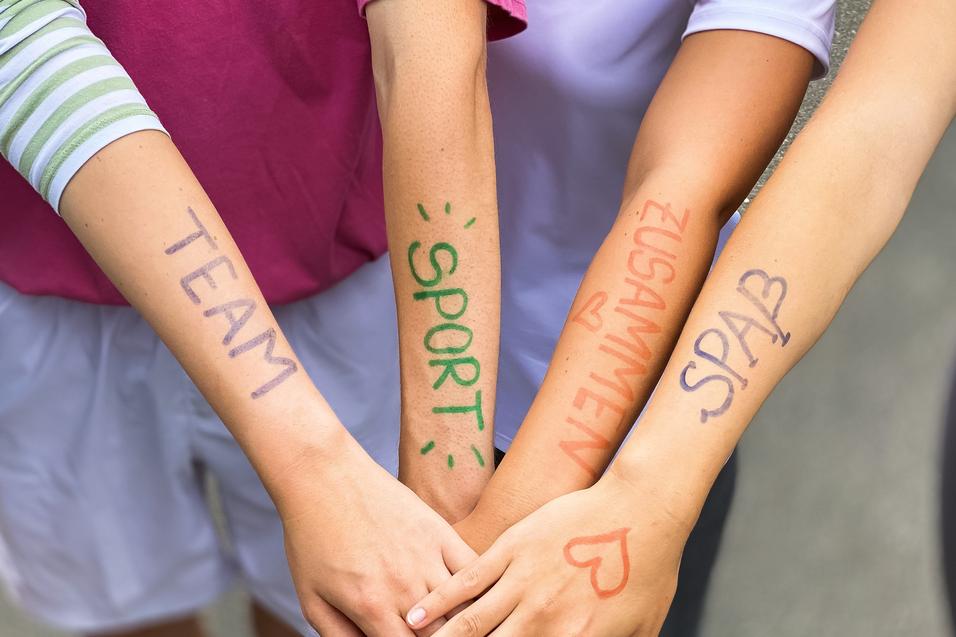 Vier Arme liegen übereinander, mit farbiger Schrift: „TEAM“, „SPORT“, „ZUSAMMEN“, „SPASS“ auf der Haut.