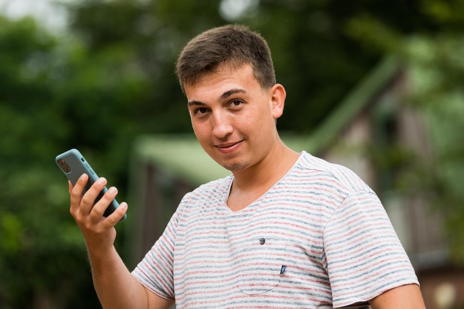 Junger Mann mit kurzen Haaren hält ein Smartphone in der Hand und schaut lächelnd in die Kamera, Hintergrund unscharf.