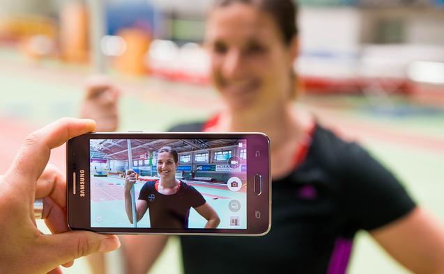 Hand hält ein Smartphone, das ein Porträt einer lächelnden Athletin mit Stab in einer Sporthalle zeigt.