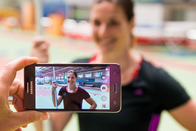 Hand hält ein Smartphone, das ein Porträt einer lächelnden Athletin mit Stab in einer Sporthalle zeigt.