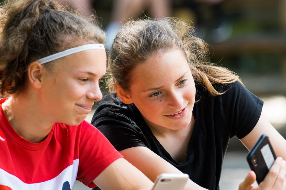 Zwei Mädchen sitzen an einem Tisch, schauen auf ihre Smartphones und lächeln dabei. Im Hintergrund sind weitere Personen sichtbar.