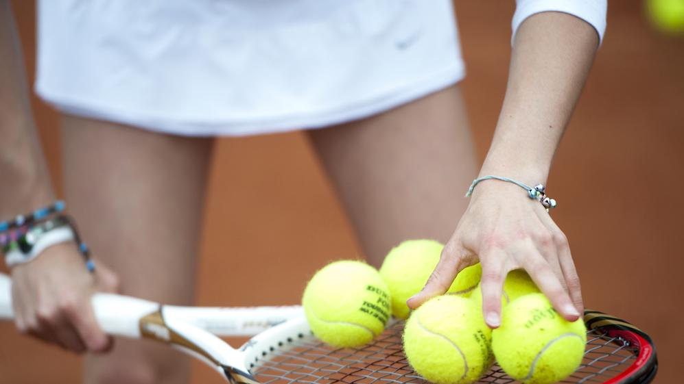 Hand greift mehrere gelbe Tennisbälle auf einem Tennisschläger, während die andere Hand die Schlägerhaltung stabilisiert.