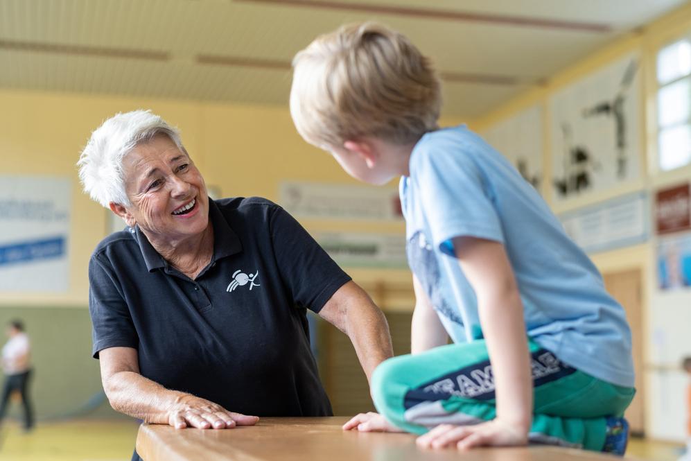 Lächelnde Trainerin spricht mit einem kleinen Jungen, der auf einem Tisch sitzt, in einer Sporthalle.