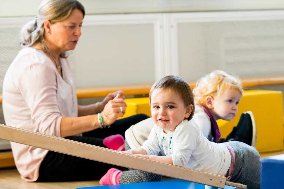 Zwei Kleinkinder spielen auf einer blauen Matte, während eine betreuende Person im Hintergrund sitzt.